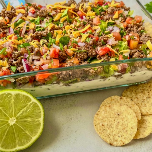 Beefy taco dip that's been freshly made and chilled served with fresh cilantro, lime, and some tortilla chips on the side