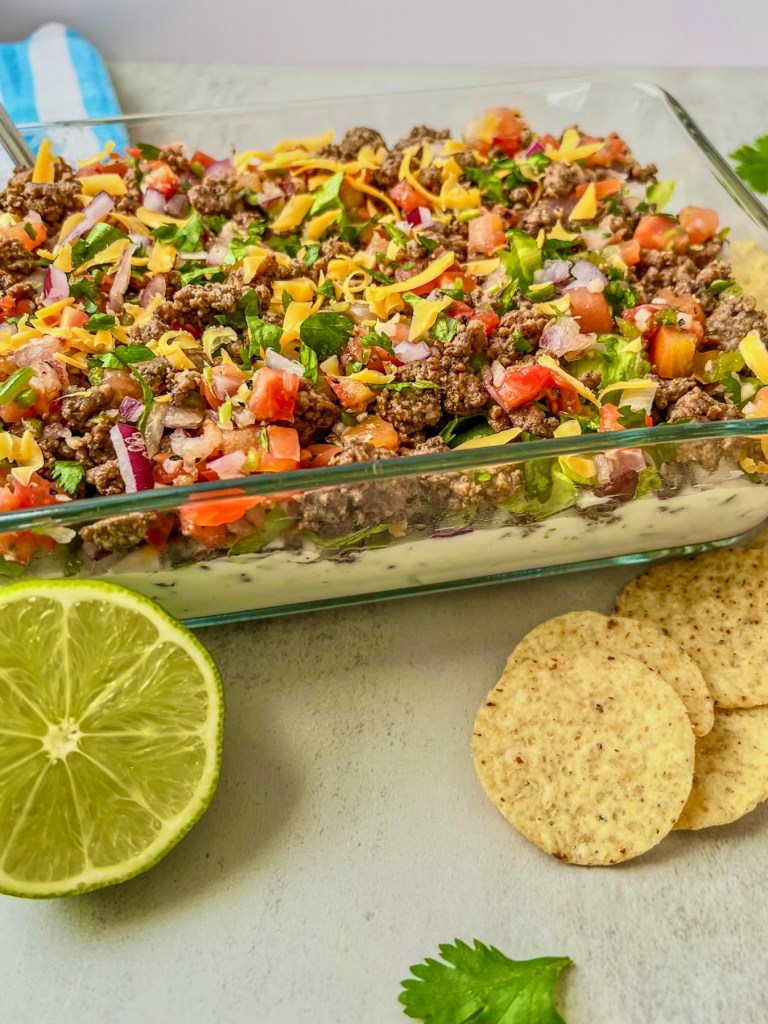 Beefy taco dip that's been freshly made and chilled served with fresh cilantro, lime, and some tortilla chips on the side