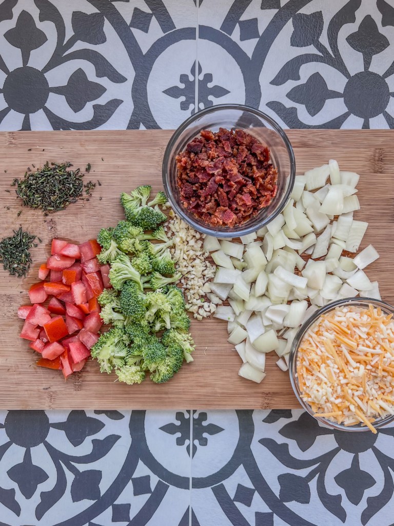 Onion, garlic, tomato, broccoli, crumbled bacon, cheese, rosemary, and thyme prepped on a cutting board