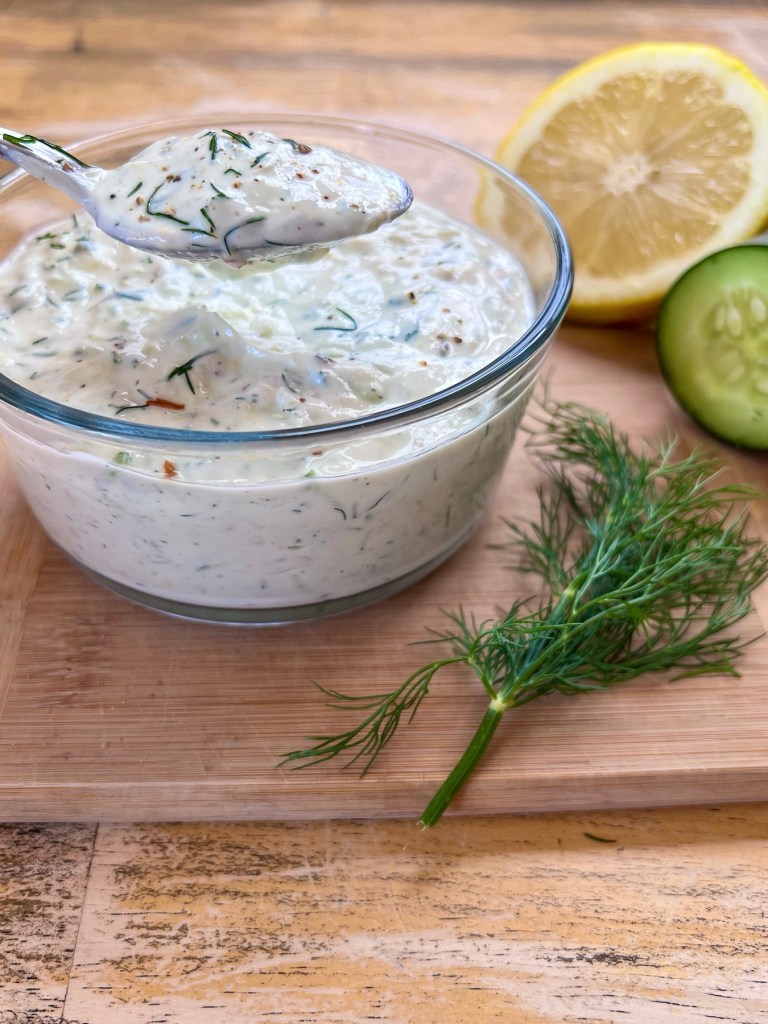 Homemade tzatziki sauce in a dish with some dill, lemon, and cucumber nearby, on a wooden cutting board