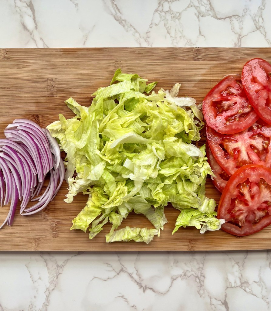 Thinly sliced red onion, a large thinly sliced tomato, and some iceberg lettuce that's been finely shredded on a wooden cutting board