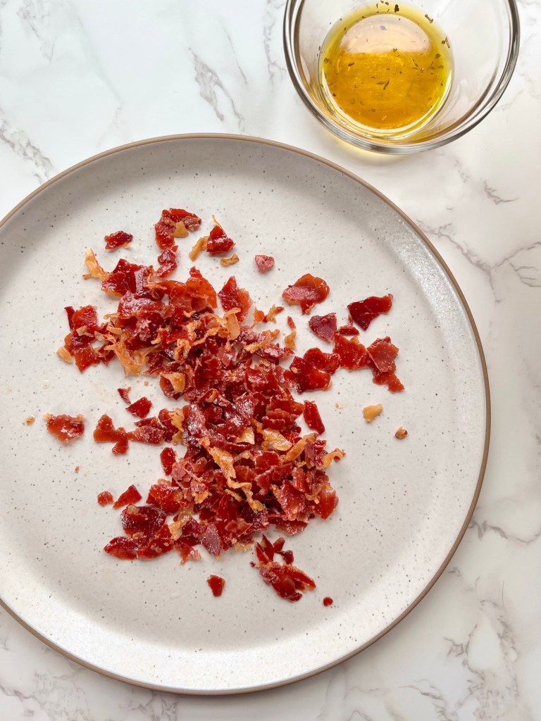 Crispy prosciutto that's been crumbled on a plate with an oil and vinegar finish in a small dish off to the side