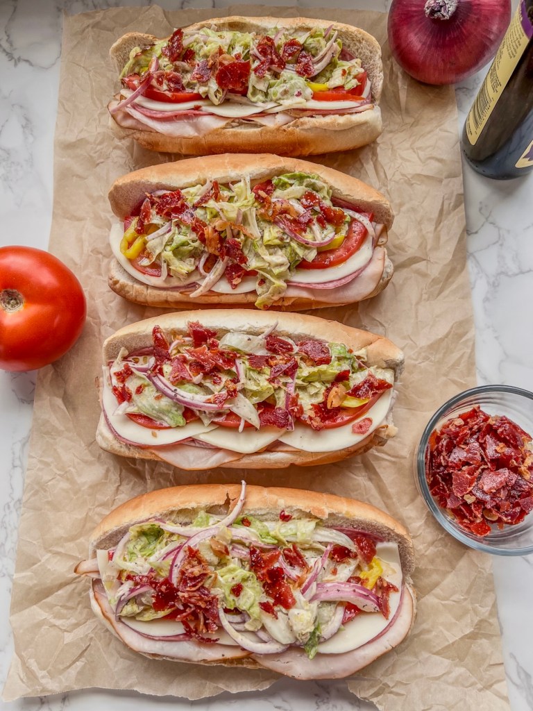 Italian grinder subs freshly assembled on some parchment paper with some tomato, red onion, red wine vinegar and crispy prosciutto off to the side
