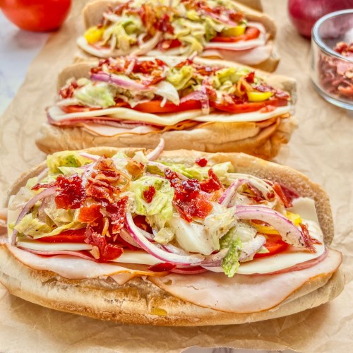 Italian grinder sub sandwiches on some parchment paper with tomato, onion, crispy prosciutto, and red wine vinegar off to the side