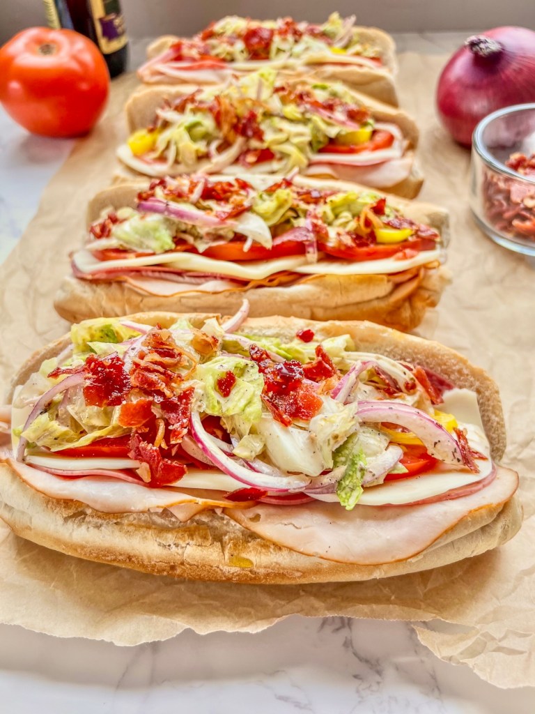 Italian grinder sub sandwiches on some parchment paper with tomato, onion, crispy prosciutto, and red wine vinegar off to the side