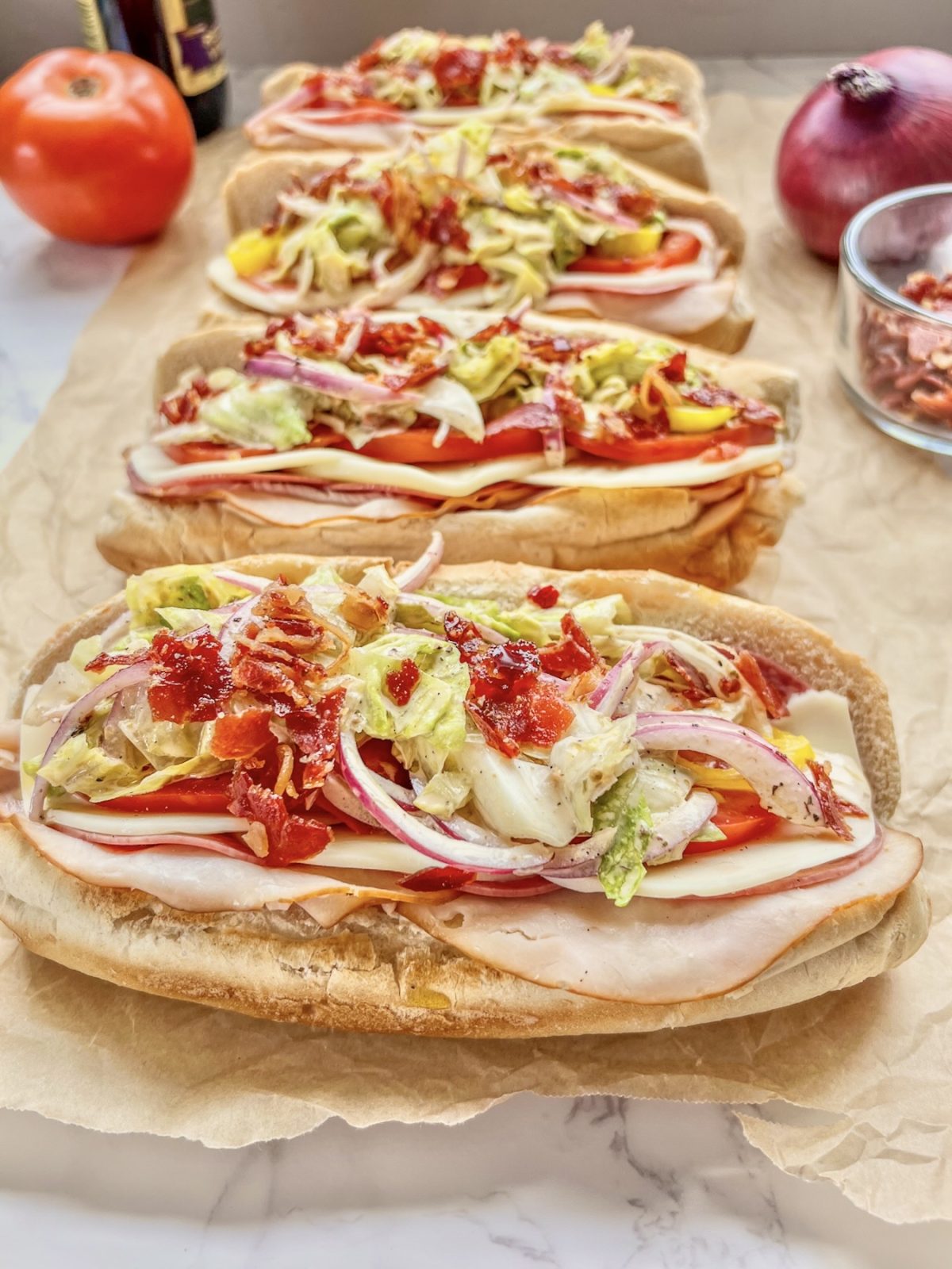 Italian grinder sub sandwiches on some parchment paper with tomato, onion, crispy prosciutto, and red wine vinegar off to the side