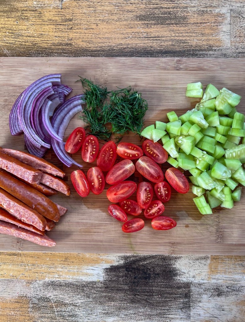 Red onion, grape tomatoes, cucumber, dill, and smoked andouille sausage cut up on a cutting board