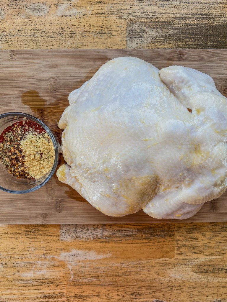 A whole raw chicken on a cutting board with a dish filled with spices for a rub next to it
