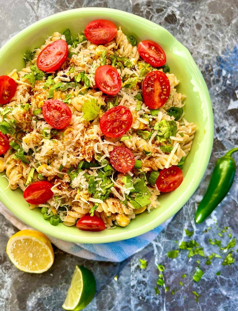 Street corn pasta salad in a bowl with some lemon, lime, jalapeño, and herbs scattered around