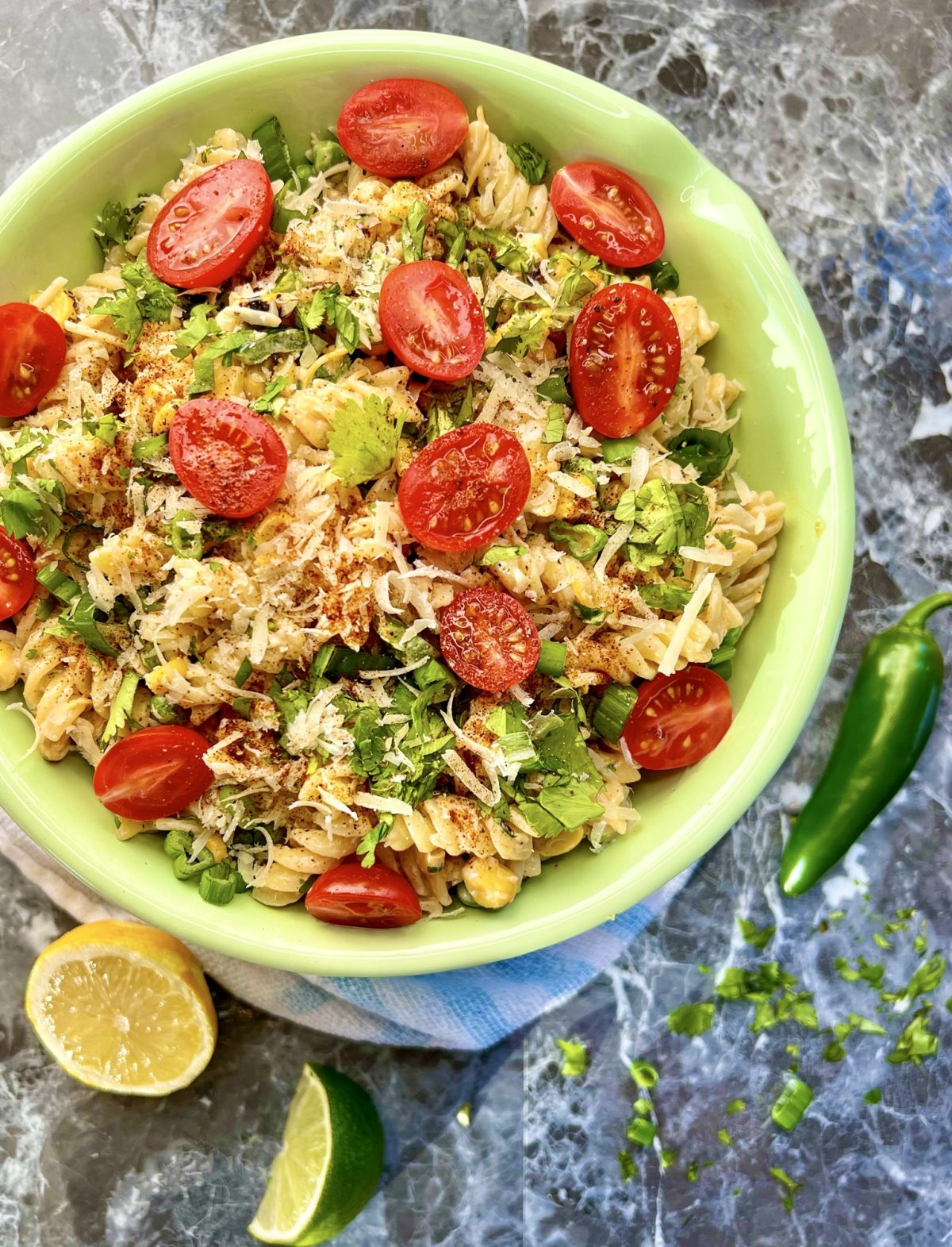 Street corn pasta salad in a bowl with some lemon, lime, jalapeño, and herbs scattered around