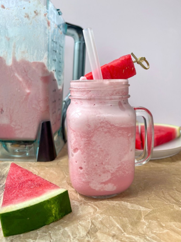 Watermelon Cloud Slushie in a glass with a blender filled with more slushie mix behind and some watermelon triangles off to the side
