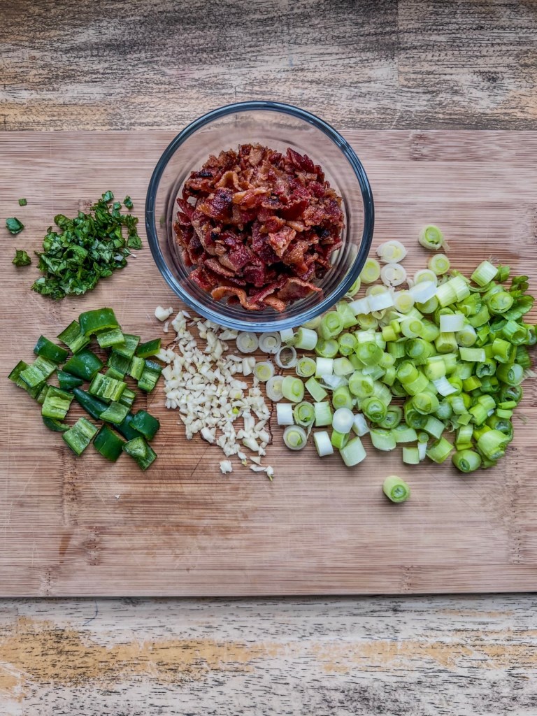 Serrano peppers, green onions, garlic, crumbled bacon, and minced cilantro prepped on a cutting board