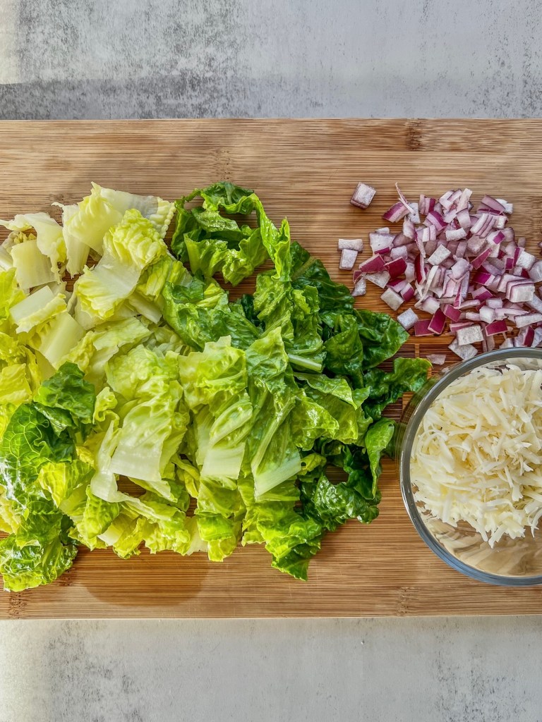 Chopped romaine lettuce, shredded parmesan cheese, and finely diced red onion on a cutting board