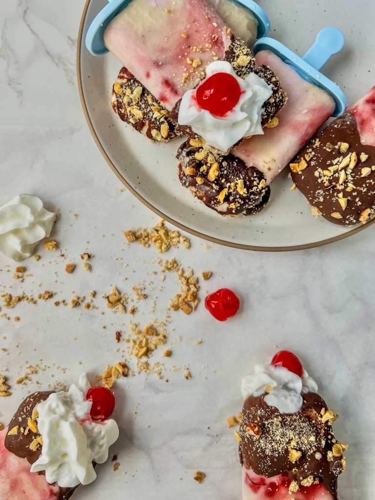 Banana split frozen yogurt pops stacked on a plate with whipped cream and maraschino cherries on top