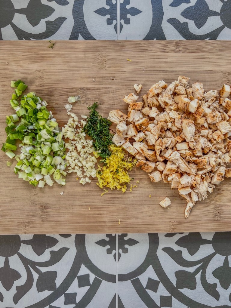 Chopped chicken, lemon zest, green onion, dill, and garlic prepped on a cutting board