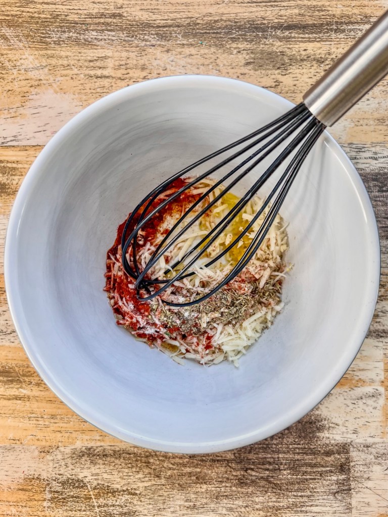 Spices, olive oil, garlic, lemon zest, and shredded parmesan cheese in a large mixing bowl with a whisk inside