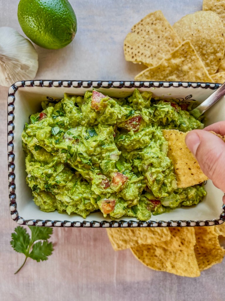Easy guacamole dip in a serving dish with some lime and garlic off to the side and a hand scooping some out with a tortilla chip