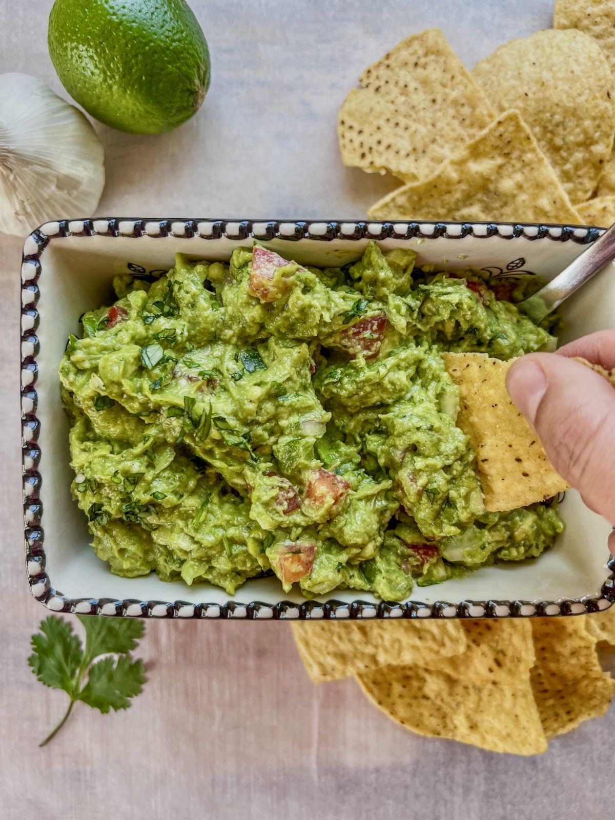 Easy guacamole dip in a serving dish with some lime and garlic off to the side and a hand scooping some out with a tortilla chip