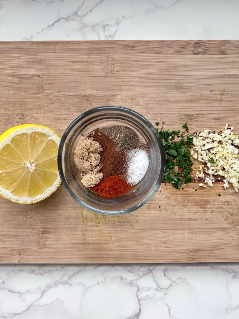 Lemon, parsley, garlic, and a small dish with a variety of spices on a cutting board