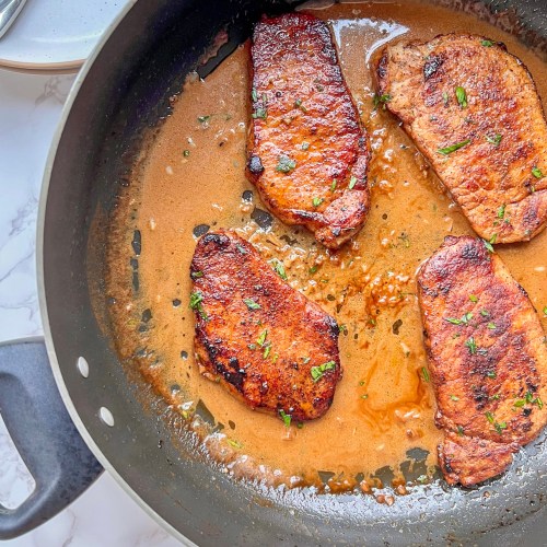 Espresso rubbed pork chops with a creamy garlic sauce in a skillet with some plates and silverware nearby