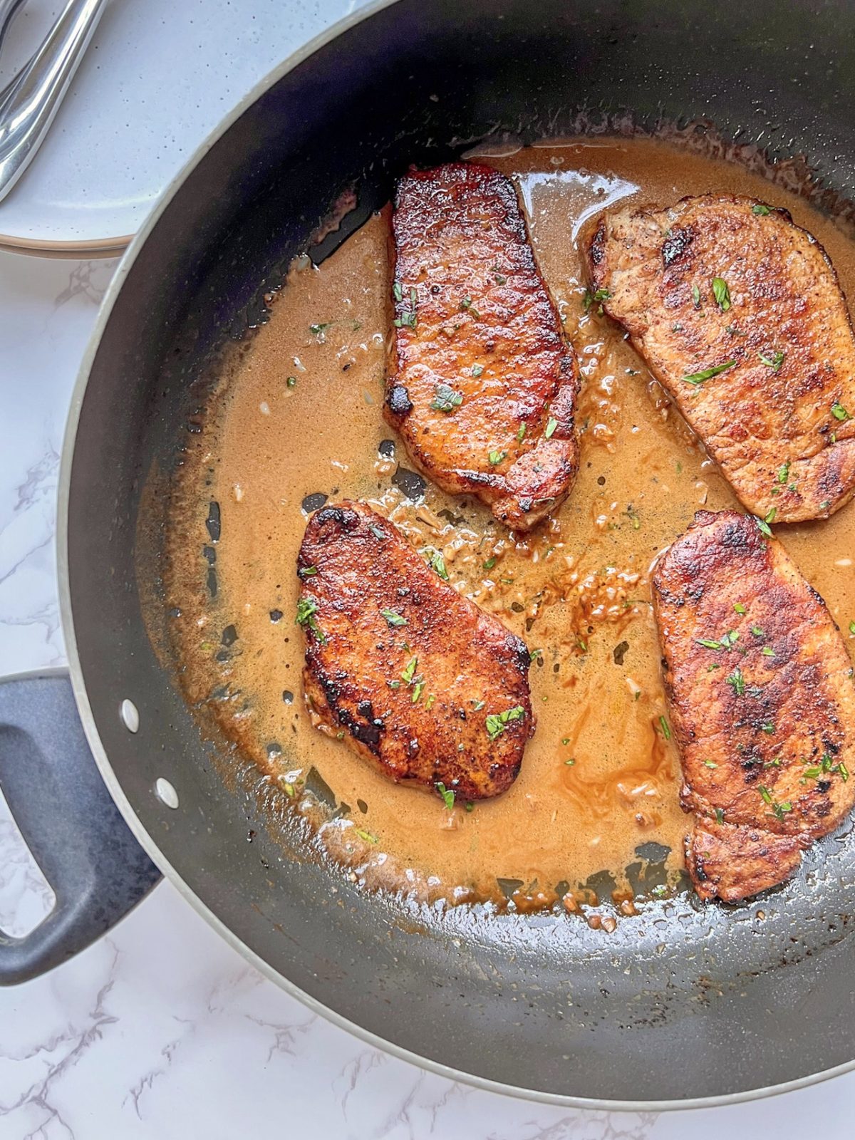 Espresso rubbed pork chops with a creamy garlic sauce in a skillet with some plates and silverware nearby