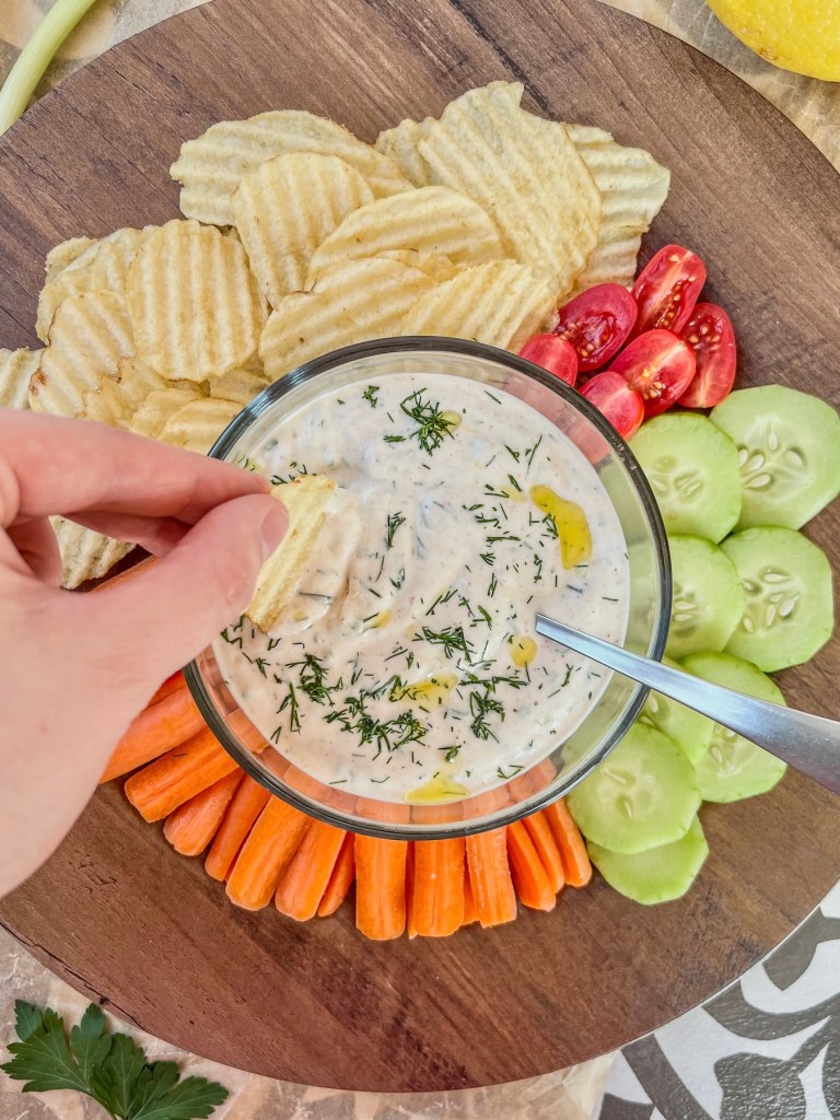 Lemon herb chip and veggie dip being scooped up with some chips with some veggies off to the side