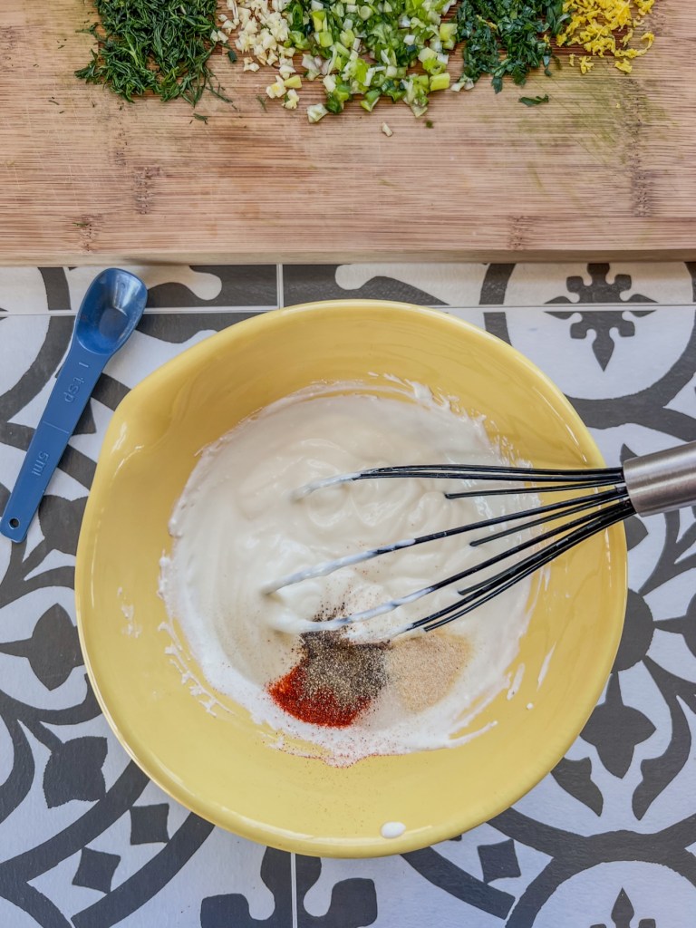 Sour cream, Greek yogurt, mayo, and spices being whisked in a bowl with some fresh herbs off to the side