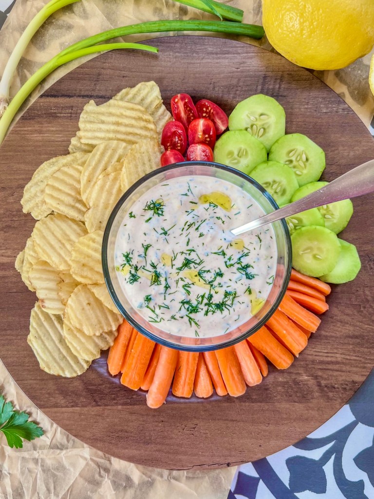Lemon herb chip and veggie dip in a small bowl with a platter filled with veggies and chips surrounding it