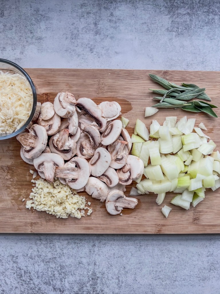 Sliced mushrooms, diced onion, minced garlic, shredded parmesan cheese, and sage leaves on a cutting board