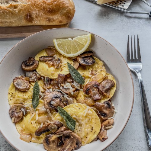Ravioli with brown butter mushrooms and sage in a bowl with a lemon wedge and a crispy baguette on the side