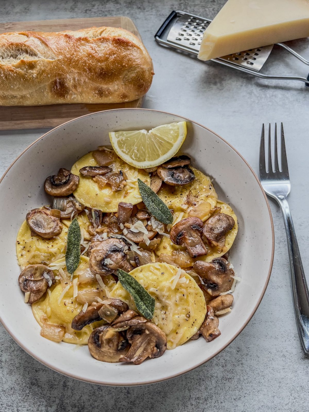 Ravioli with brown butter mushrooms and sage in a bowl with a lemon wedge and a crispy baguette on the side