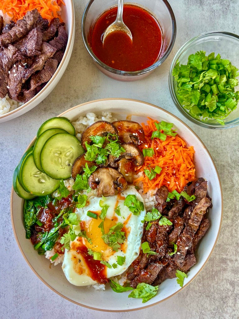 Steak bibimbap in a bowl with cucumber, carrots, spinach, green onion, cilantro, a sunny side up egg, and a lemon gochujang sauce presented on top