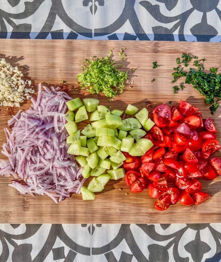 Onion, tomato, cucumber, cilantro, lime zest, and garlic prepped on a cutting board