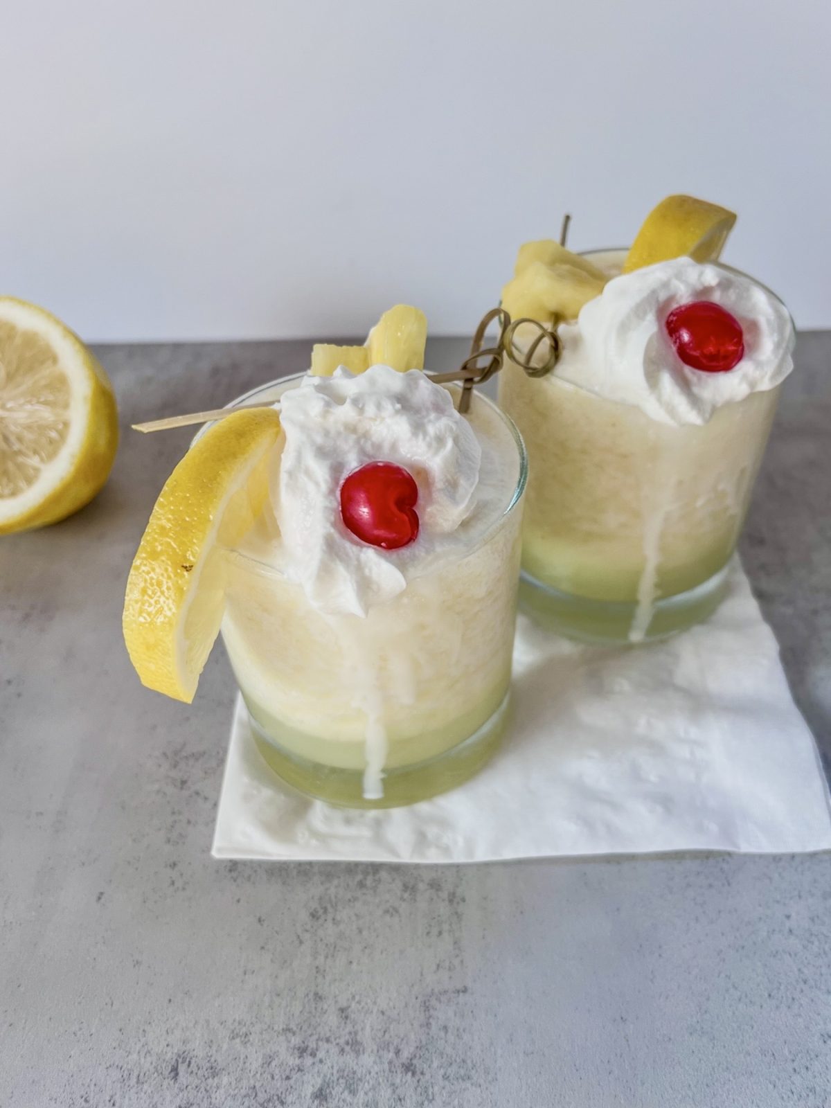Two frozen pineapple creamsicle colada drinks with whipped cream, cherries, pineapple, and lemon wedges for garnish