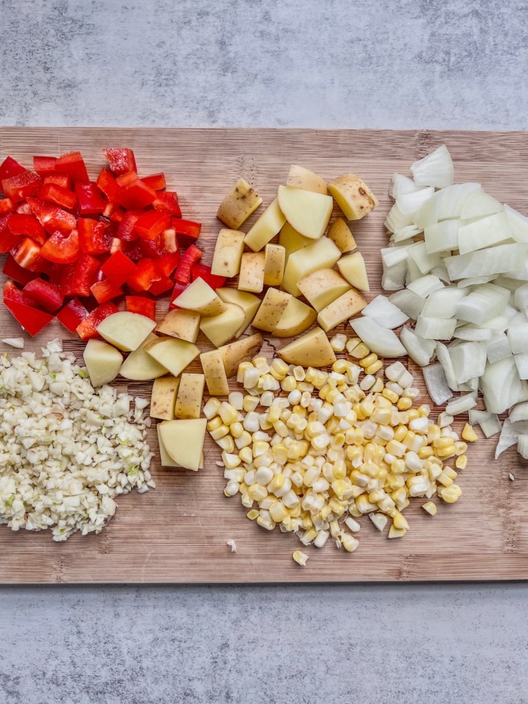 Onion, garlic, red bell pepper, potatoes, and corn prepped on a cutting board