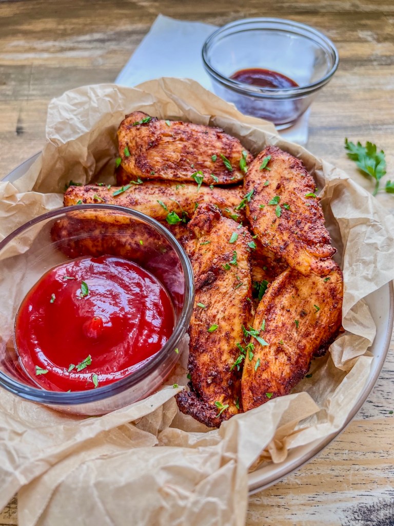 Homemade air fryer chicken tenders on a white plate with a side of ketchup and bbq sauce