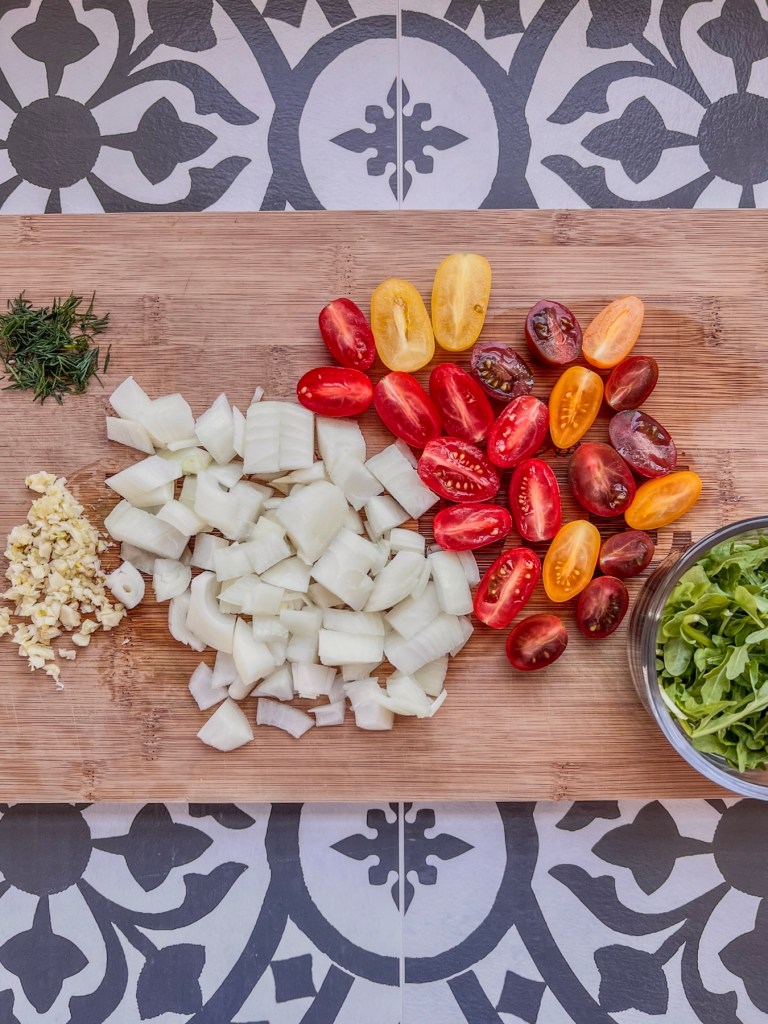 Grape medley tomatoes, garlic, yellow onion, dill, and arugula prepped on a cutting board