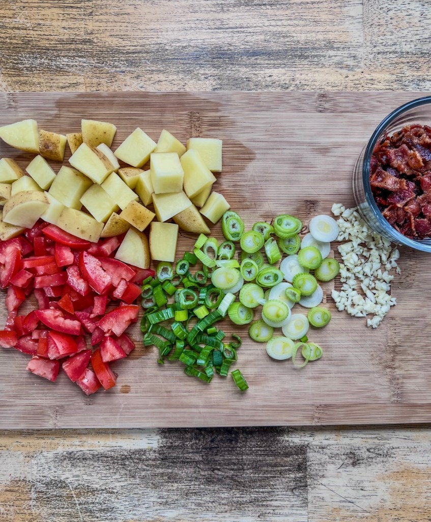 Potatoes, tomatoes, green onion, garlic, and bacon prepped on a cutting board.