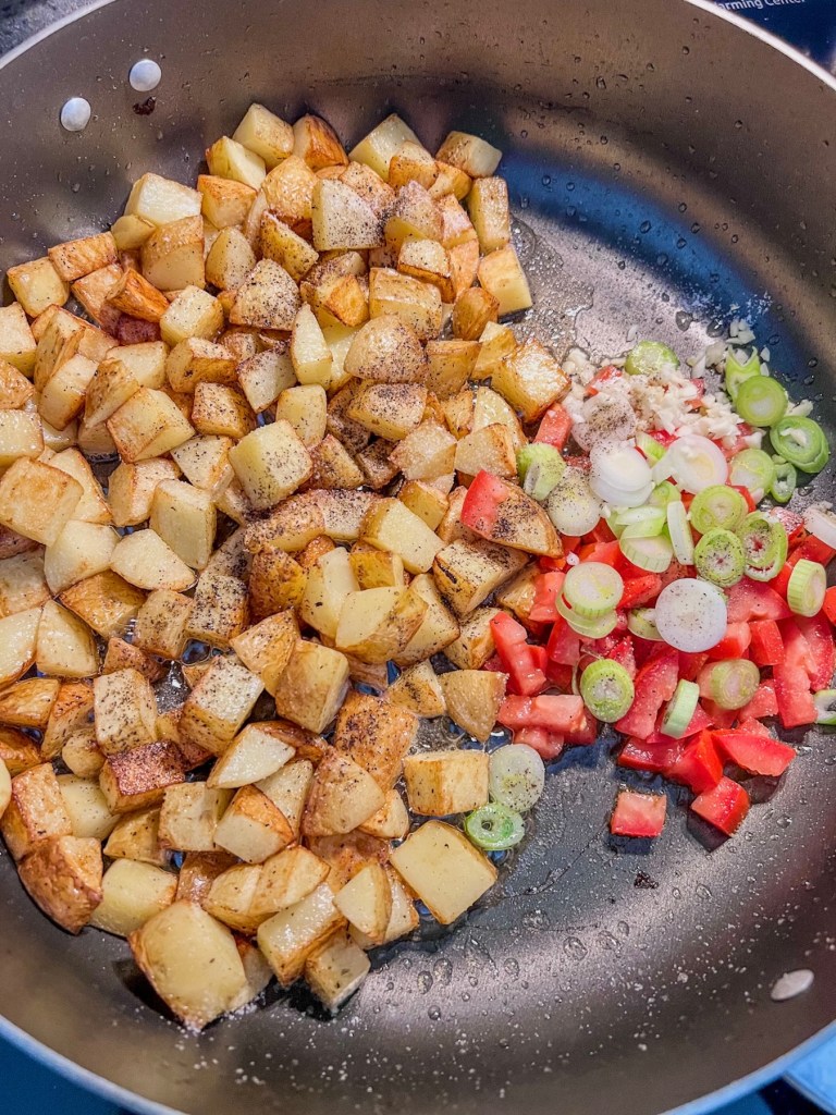 Crispy yellow potatoes in a skillet with some garlic, green onion, and tomato cooking on the side
