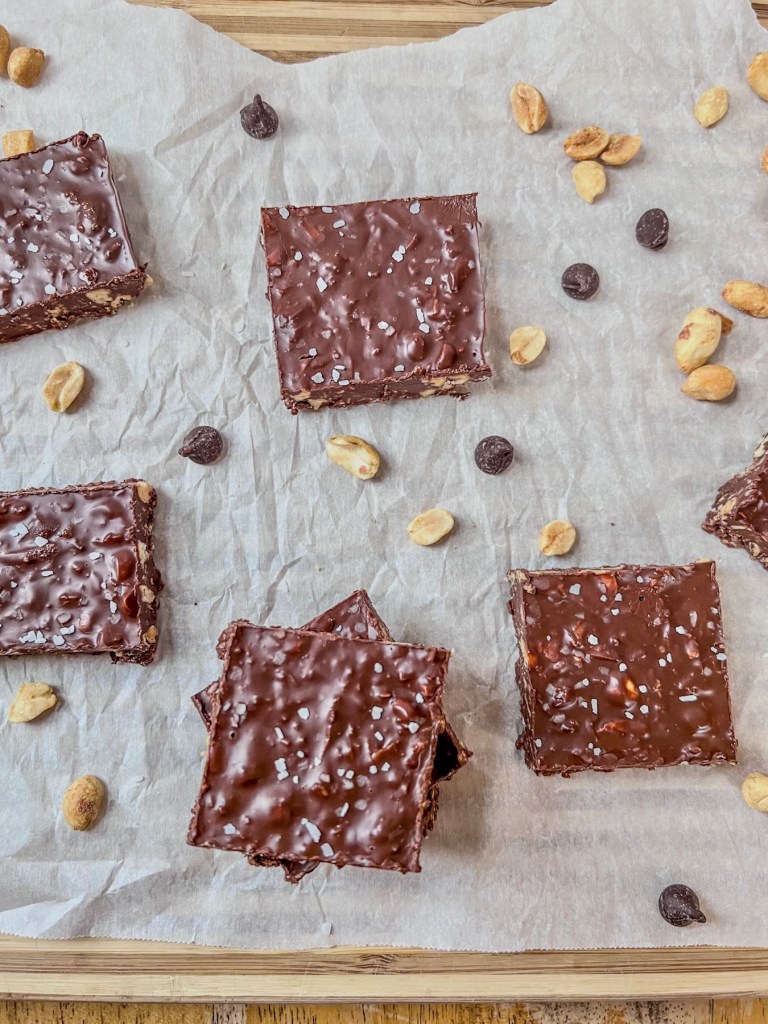 Freshly made chocolate bars with some chocolate chips and roasted peanuts scattered around them on parchment paper.