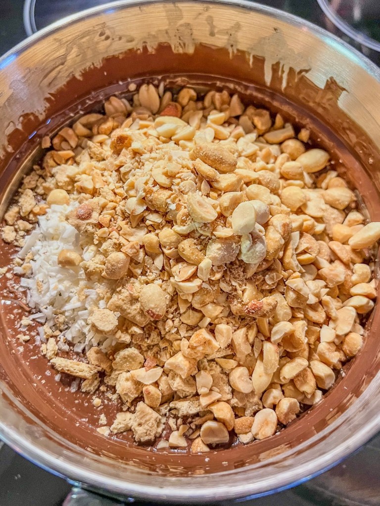 Melted chocolate chips with some peanuts, coconut flakes, and crushed graham crackers ready to be stirred in.