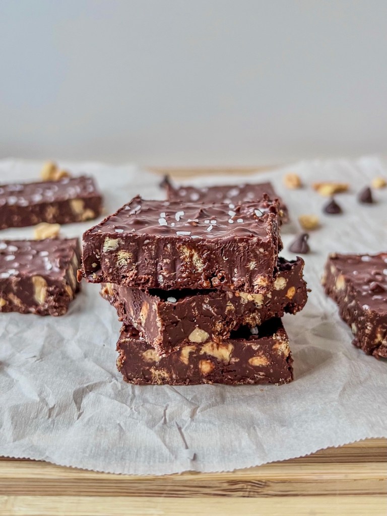 Coconut crunch chocolate squares stacked up on some parchment paper with some peanuts and chocolate chips scattered around