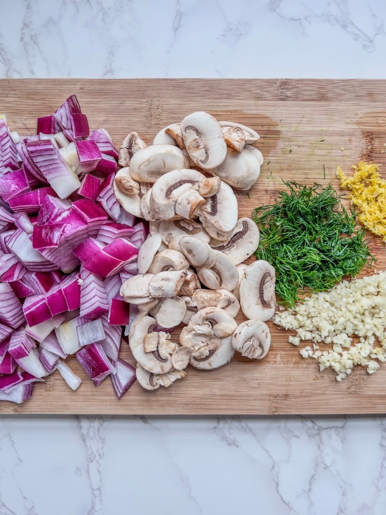 Diced red onion, minced garlic, sliced mushrooms, chopped dill, and lemon zest prepped on a cutting board