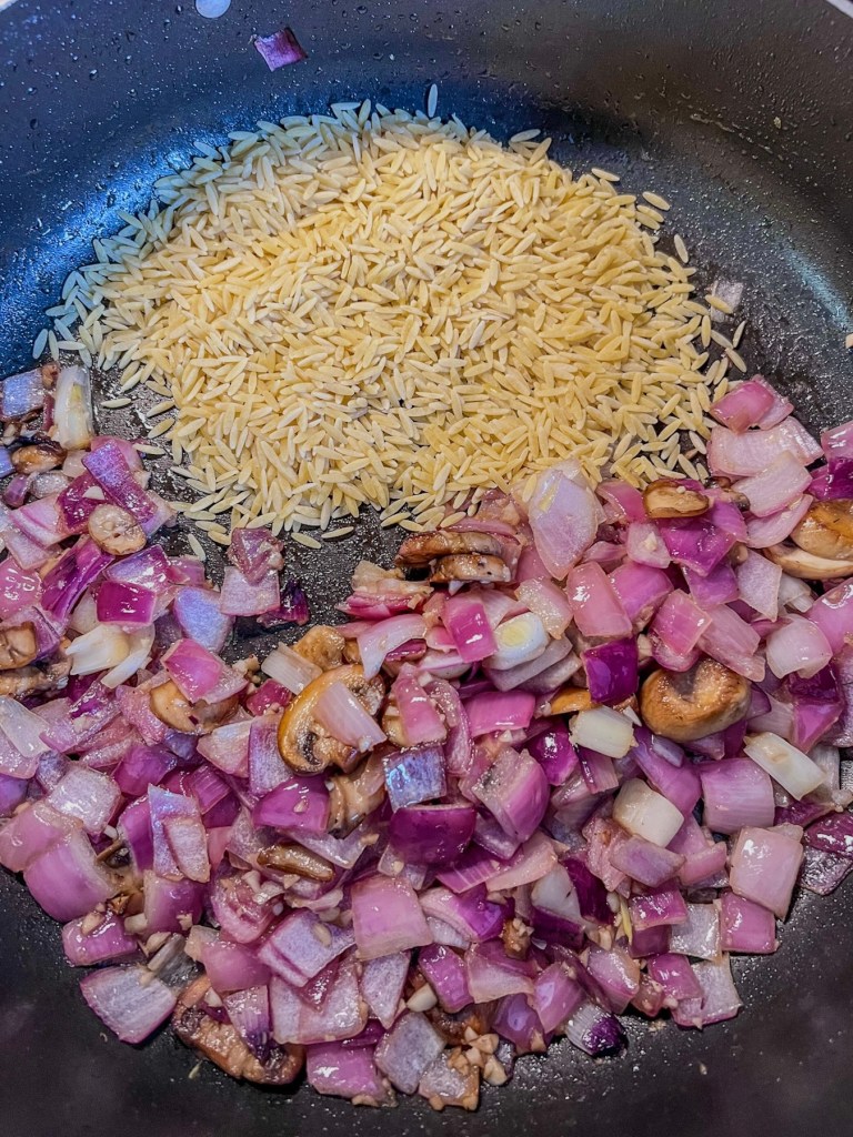 Red onion, mushroom, garlic, and orzo cooking in a skillet on the stove
