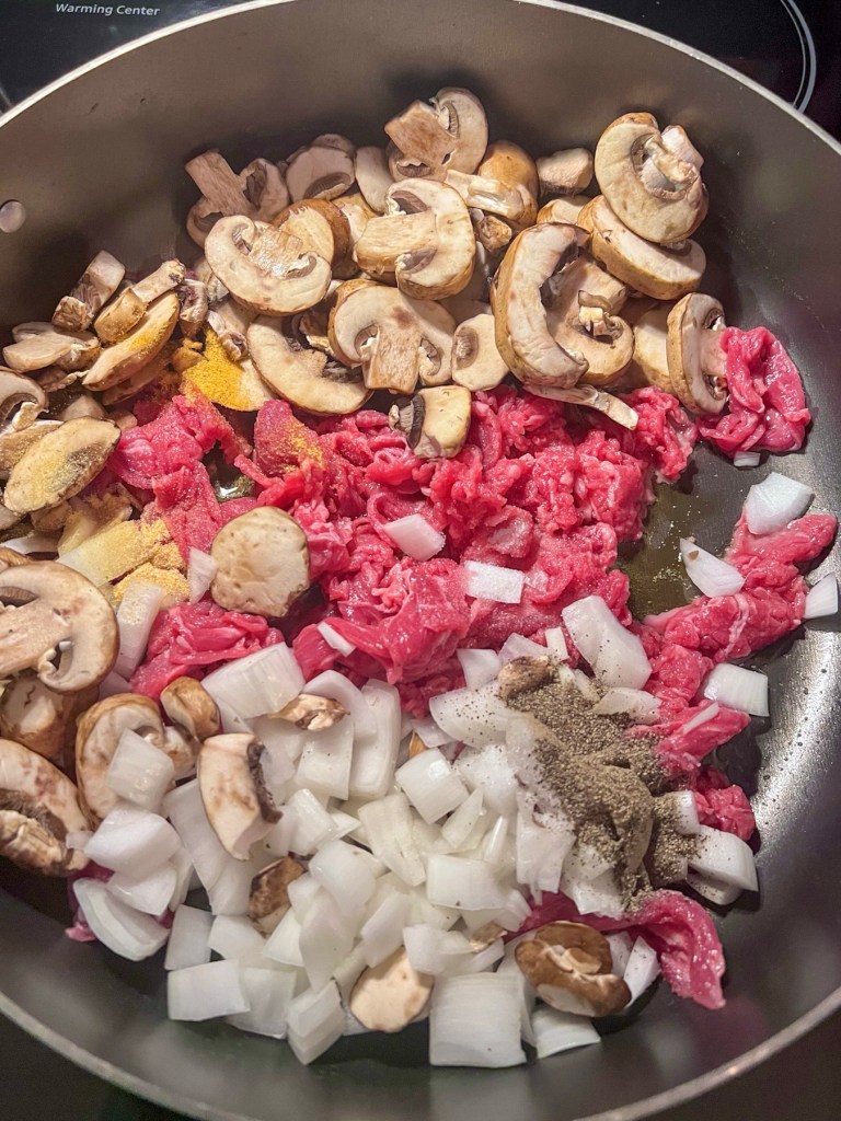 Mushrooms, onion, steak, spices, and oil cooking in a skillet on the stove.