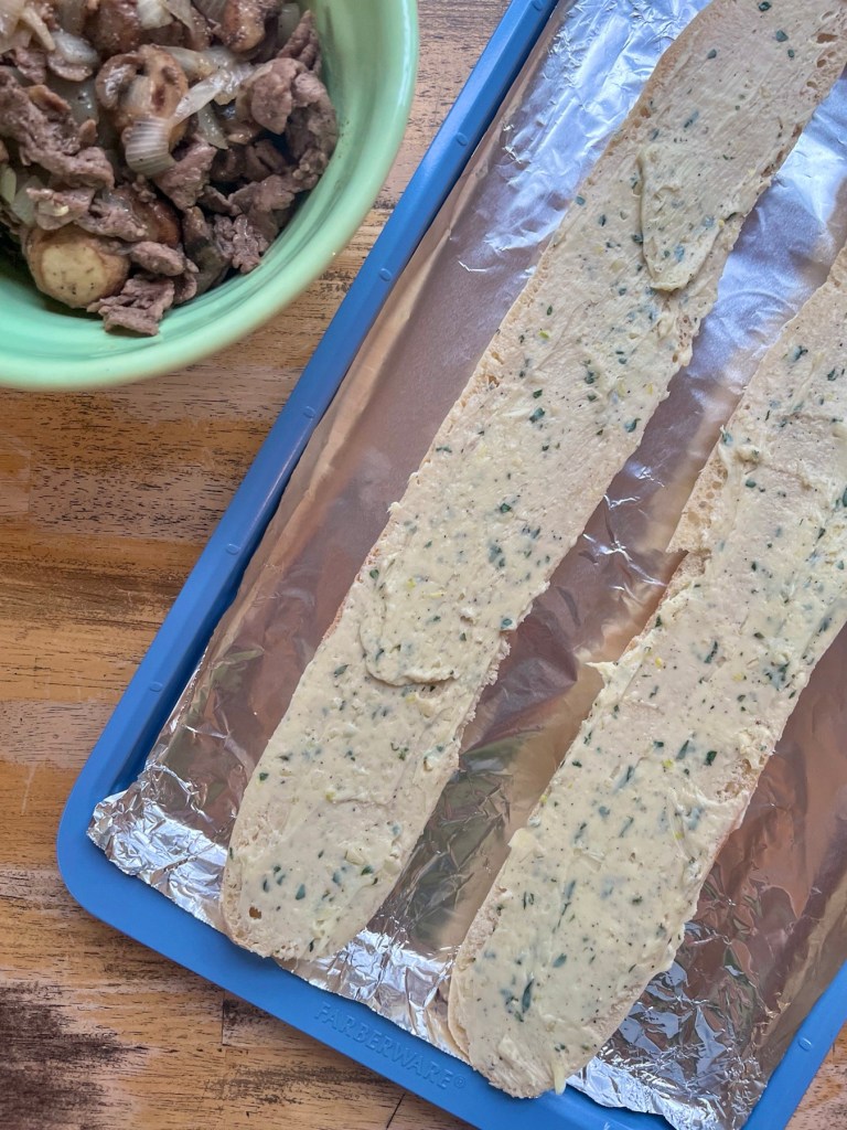 A baguette that's been sliced in half with herb butter spread on it and a steak filling mixture off to the side