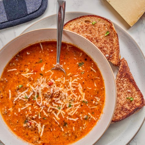 Tomato basil sausage soup in a bowl with some grilled cheese and a parmesan wedge off to the side.