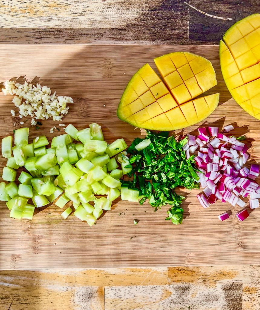 Mango, cilantro, cucumber, red onion, and garlic prepped on a cutting board