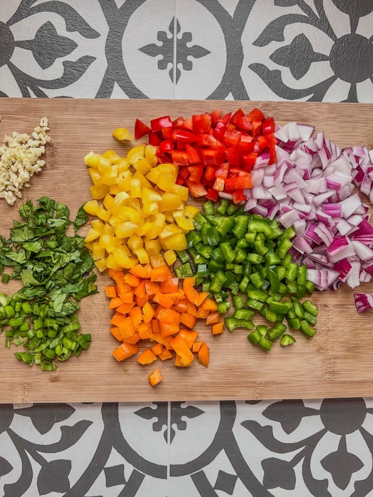 Bell pepper, red and green onion, garlic, and cilantro chopped up on a wooden cutting board.