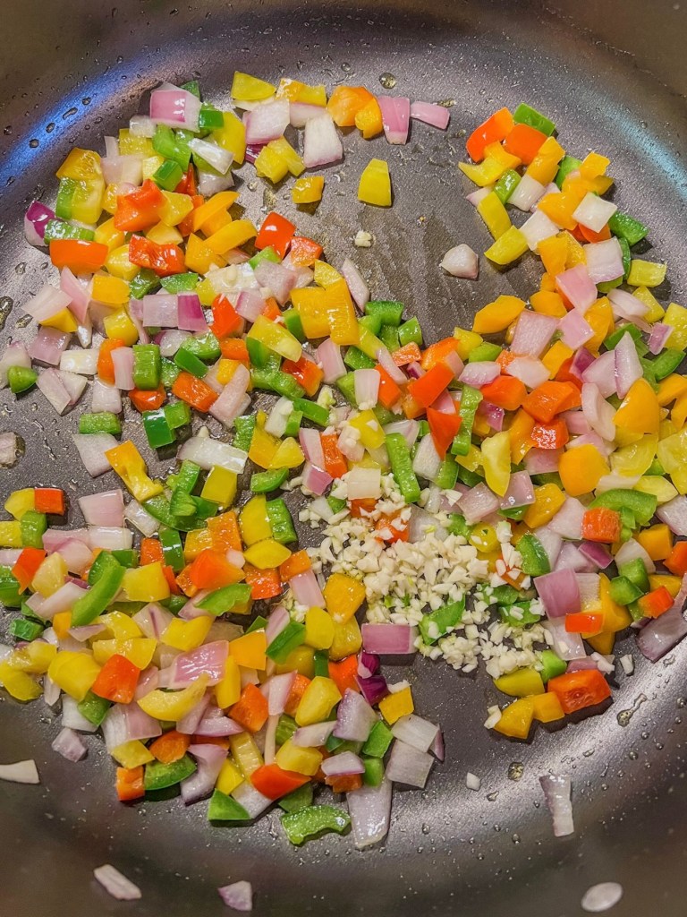Bell peppers, onions, jalapeño, and garlic cooking in a large skillet on the stove.
